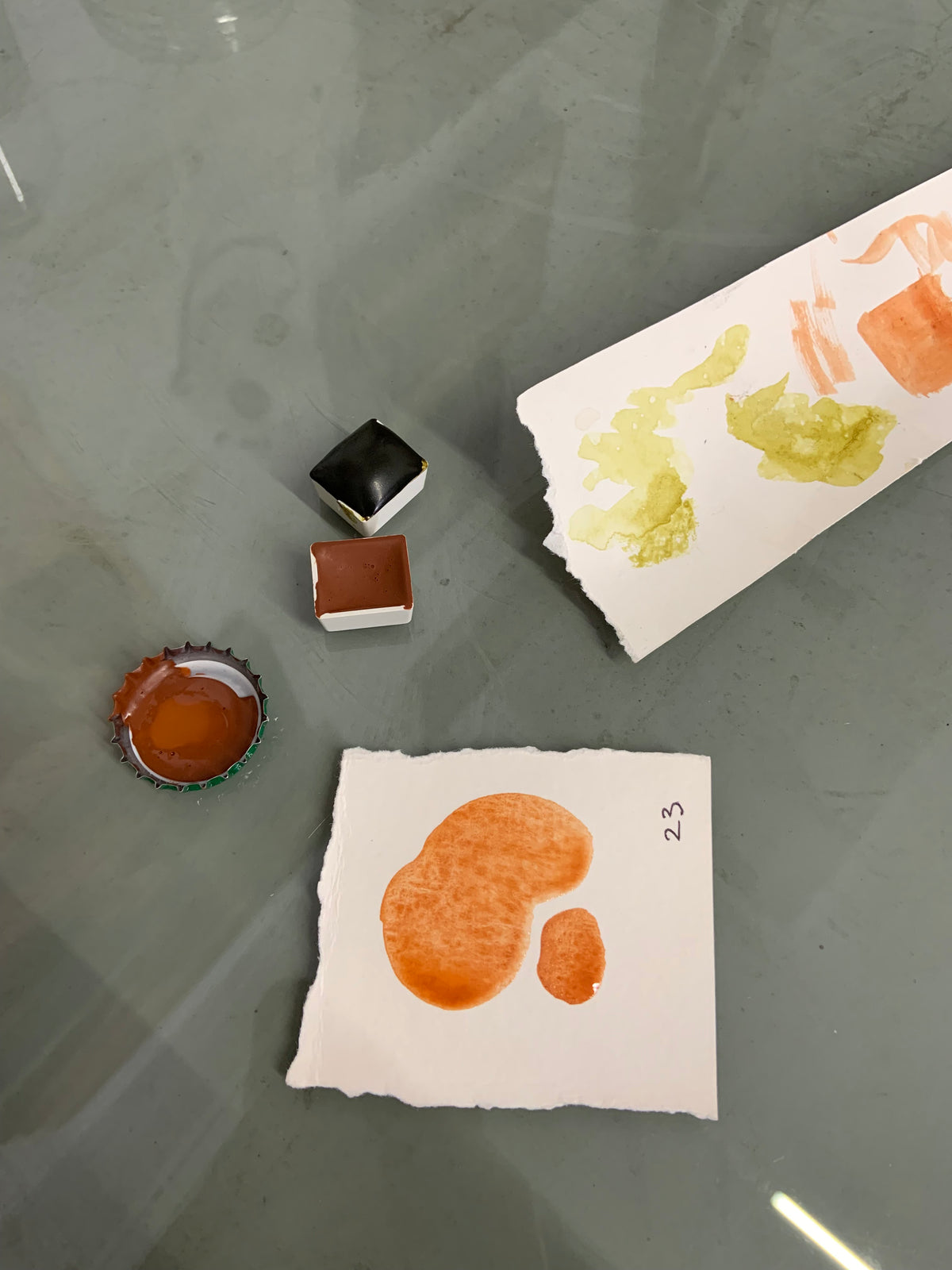 Two sheets of textured white paper with orange and yellow paint swatches, alongside two small paint cubes and a bottle cap filled with orange paint, arranged on a gray reflective surface.
