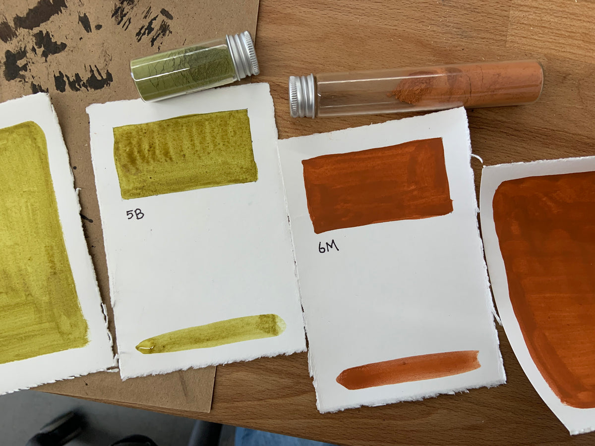Four handmade watercolor paper swatch cards displaying paint samples in olive green, ochre yellow, burnt orange, and terracotta red, arranged on a wooden table alongside two small glass pigment vials.