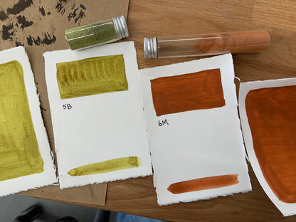 Four handmade watercolor paper swatch cards displaying paint samples in olive green, ochre yellow, burnt orange, and terracotta red, arranged on a wooden table alongside two small glass pigment vials.